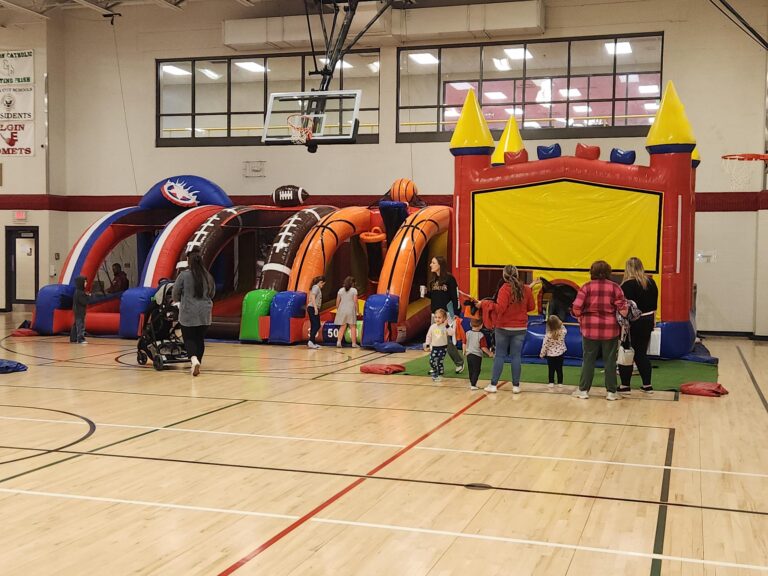 Children and families enjoying sports-themed inflatable obstacle course and bounce house inside the YMCA gymnasium at a MaxAiir event